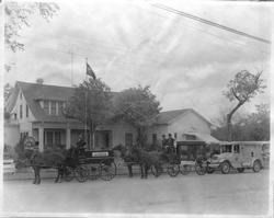 Horses and carriages parked in front of a building with an American flag. A car is also present.