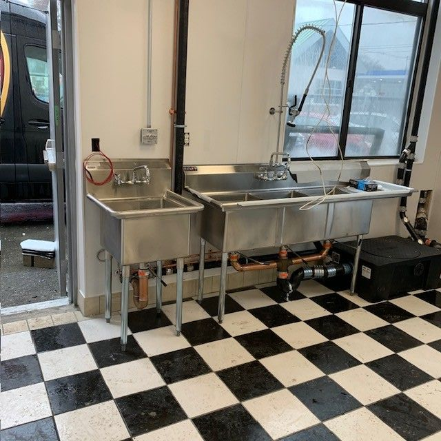 Plumber in black gloves uses a wrench to tighten plumbing under a white sink.