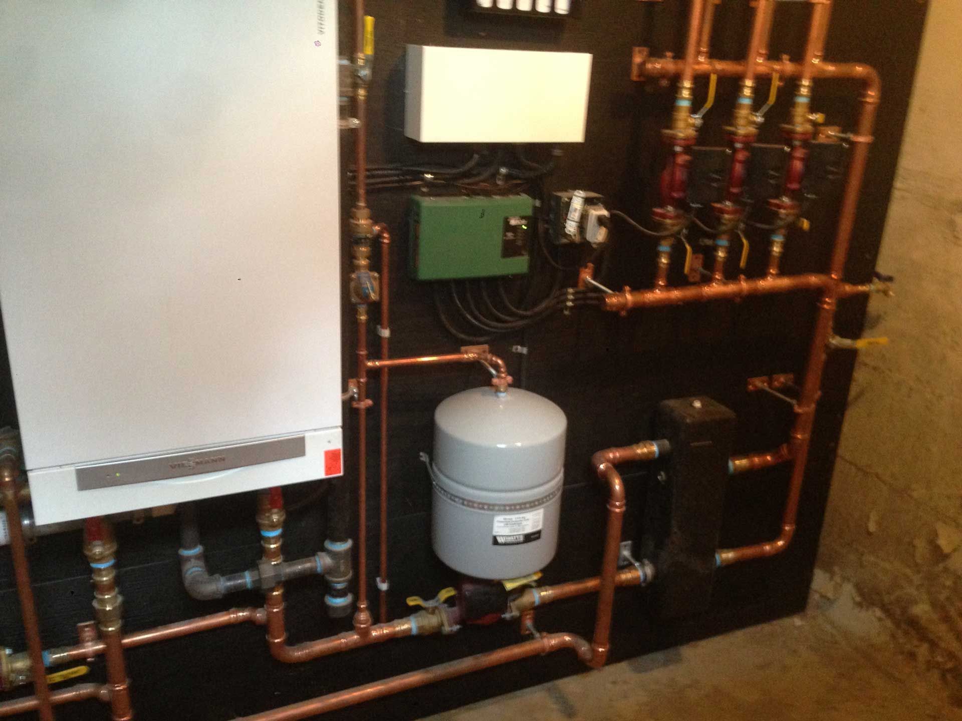 Copper pipes and heating components on a black panel; boiler, tank, pump, and valves in a basement.