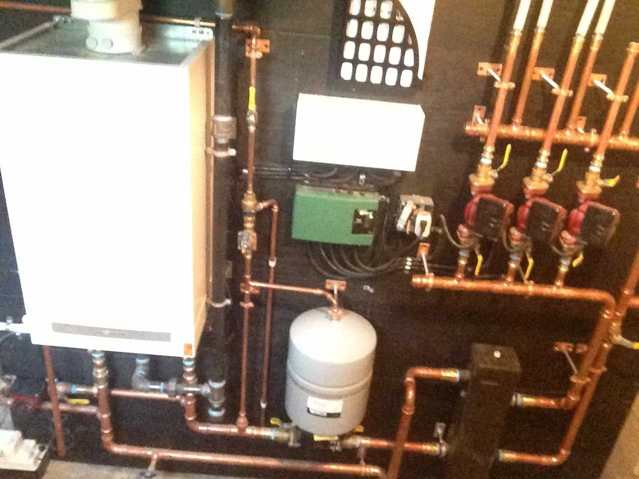 Copper pipes and tank connected to a white boiler on a black wall; heating system setup.