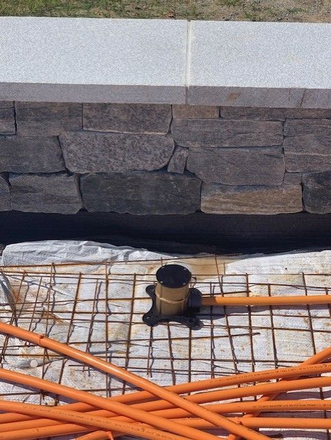 Construction site with stone wall, concrete base, rebar grid, orange conduit, and plumbing fixture.