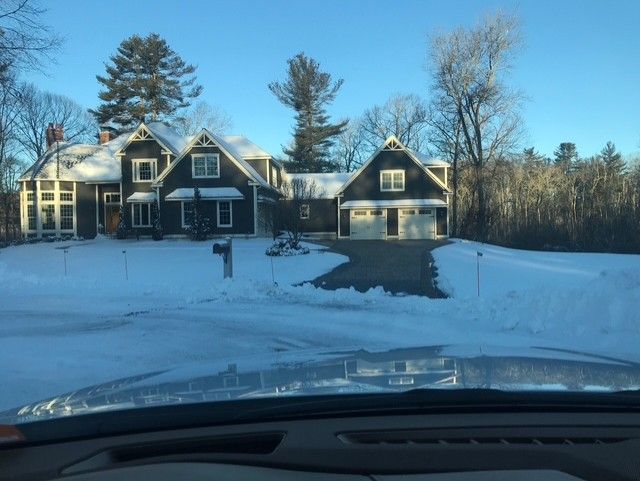 Dark house with snow-covered lawn and driveway under a clear, sunny sky. Garage is attached on the right.