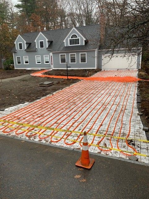 Orange heating tubes laid on a driveway of grey stone, in front of a two-story house.
