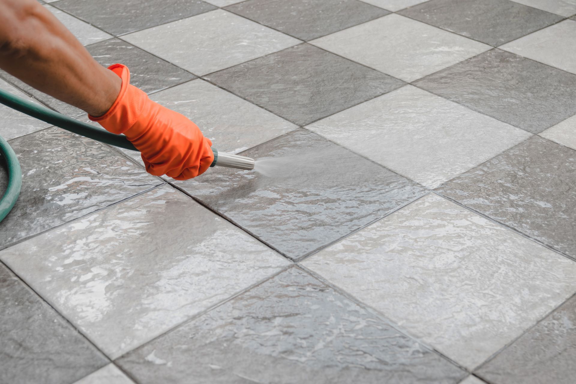 A person wearing bright orange rubber gloves is using a hose to clean the tile floor.