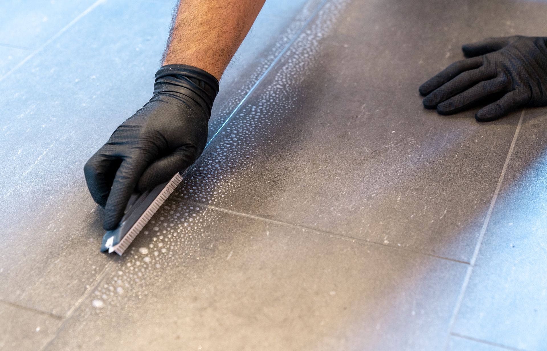 Professional cleaner meticulously scrubbing grout lines with a specialized brush and foamy soap on a sleek gray tiled bathroom floor.