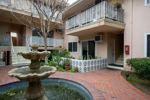 A courtyard view of an apartment complex featuring a tiered water fountain, red brick walkway, balconies, and greenery.