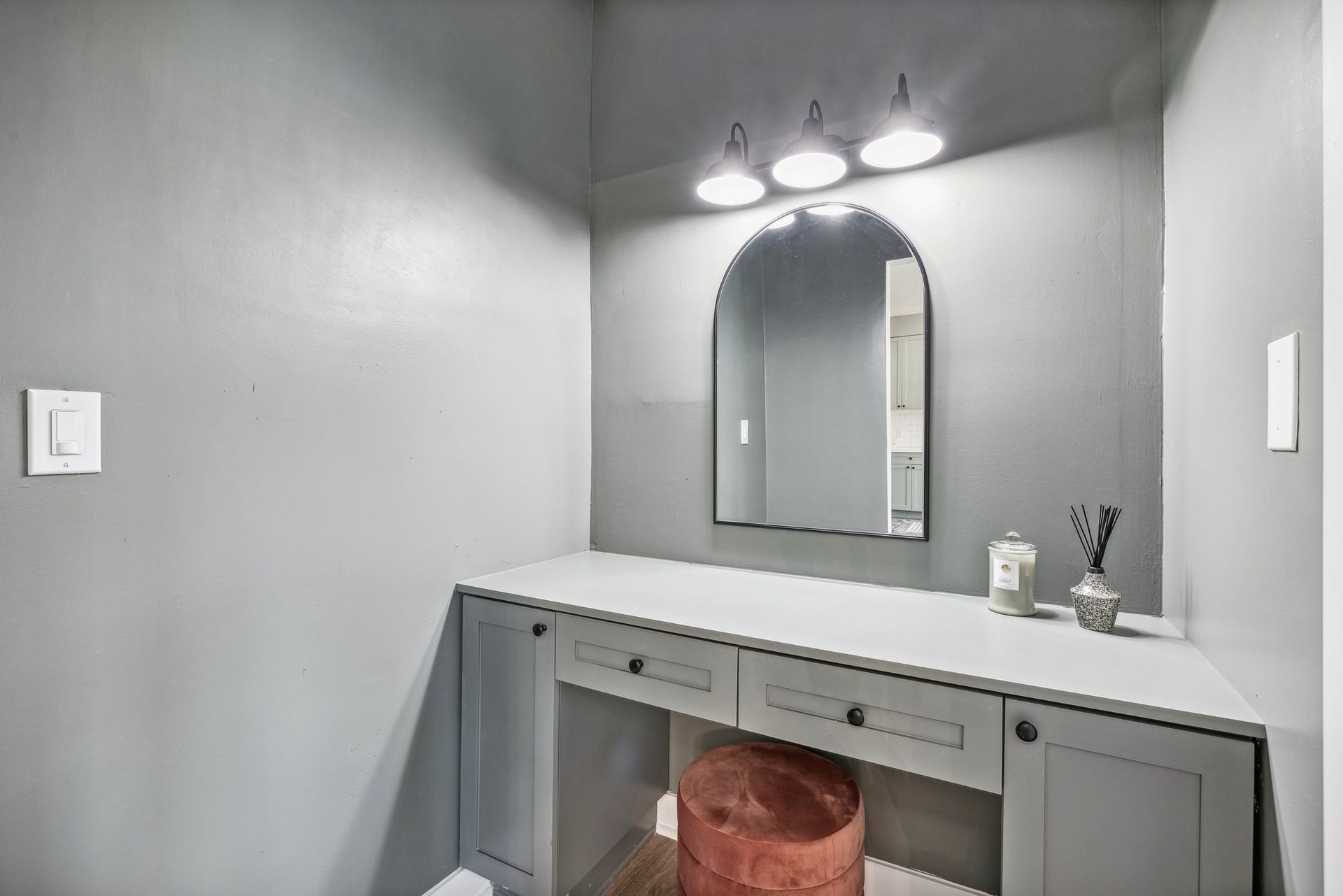 A modern gray vanity area with an arched mirror, built-in drawers, soft lighting, and a pink velvet stool for seating.