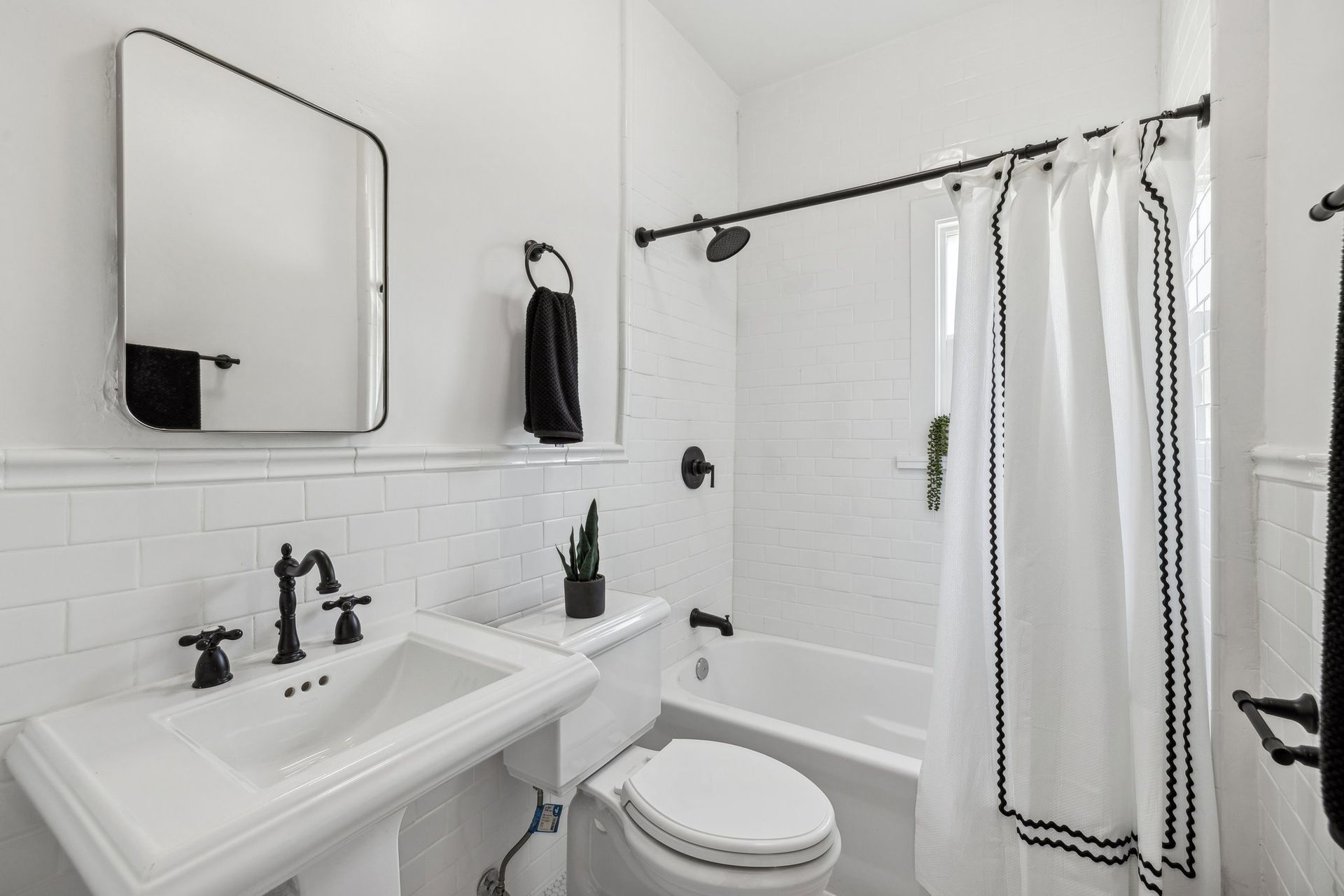 A modern white bathroom with subway tiles, a pedestal sink, black fixtures, and a shower curtain with black trim details.