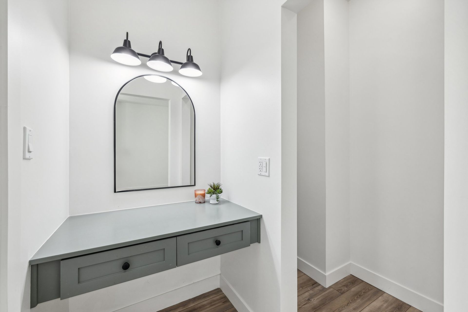 A modern vanity nook with a gray floating desk, arched mirror, black light fixtures, and minimalist decor.