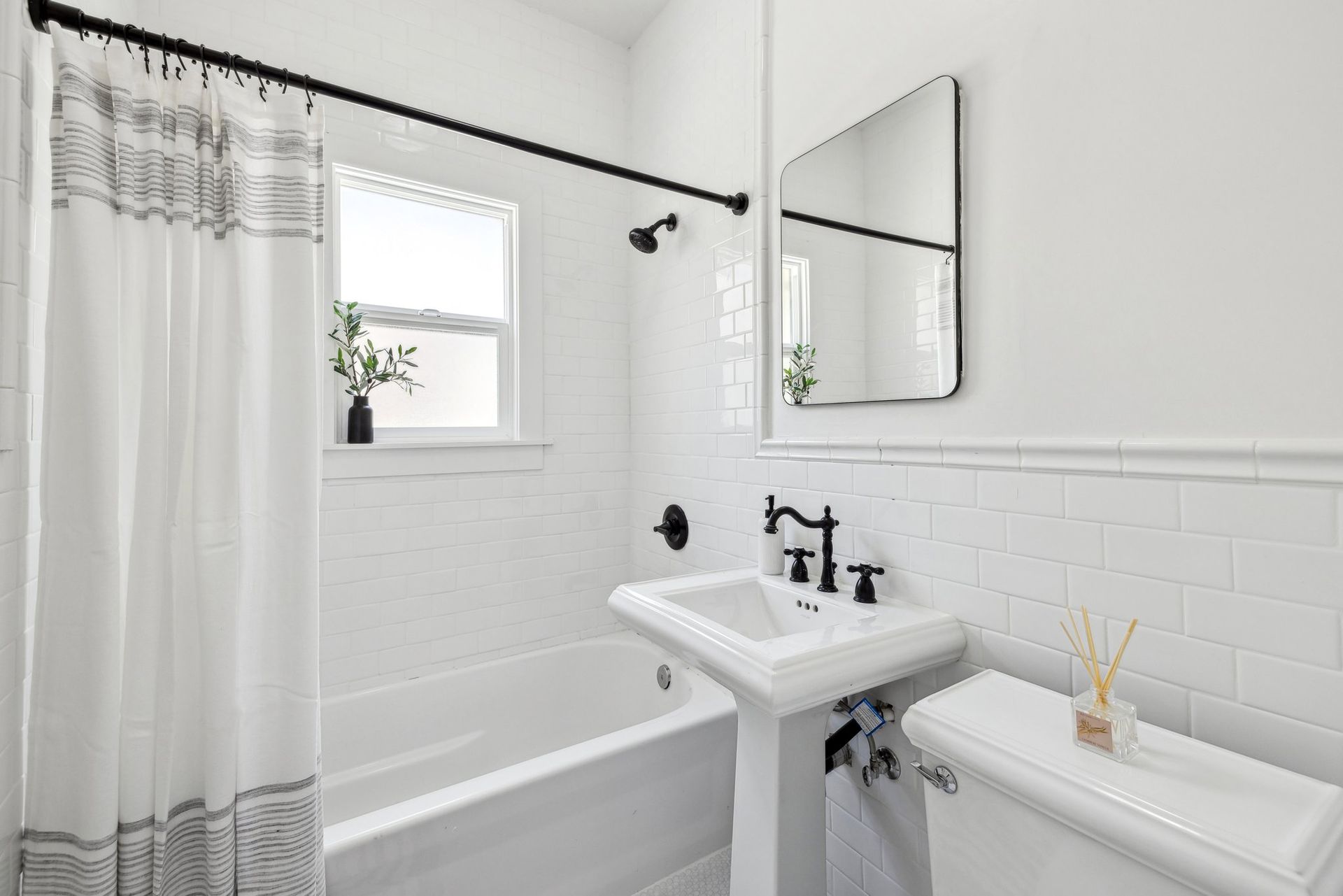 A bright, modern bathroom with white subway tiles, a pedestal sink, black fixtures, and a bathtub with shower curtain.