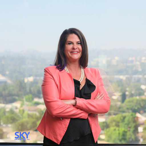 Kari Negri, CEO of Sky Properties, Inc., stands confidently in a coral blazer against a scenic city backdrop