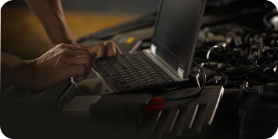 A person is typing on a laptop in front of a car engine. | Oceanside Tire and Service Center Tire Pros