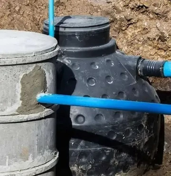 A Black Septic Tank With a Textured Surface is Connected to a Cement Chamber via Blue Pvc Pipes
