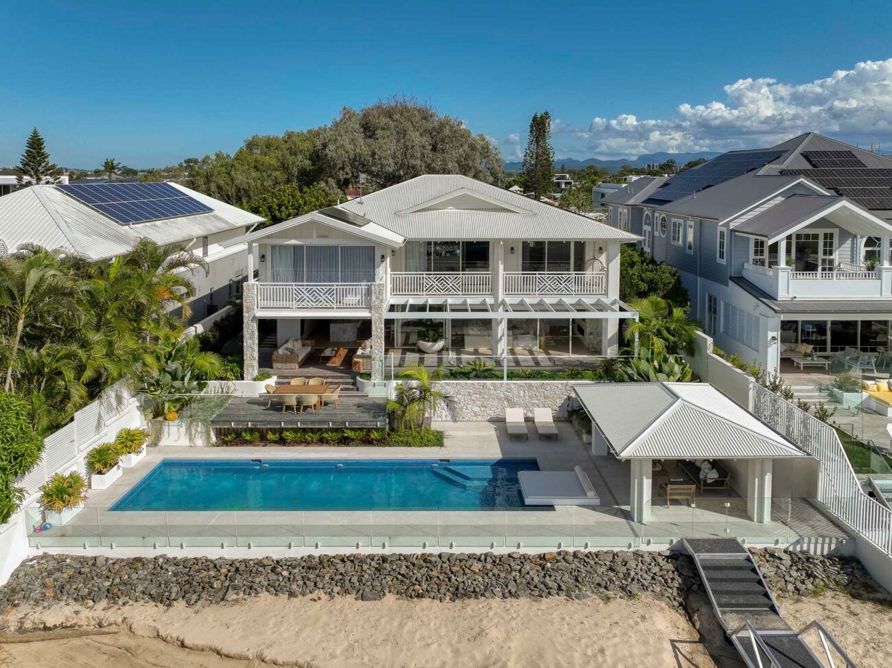 Coastal two-story white house with a pool, cabana, and dock on a sandy beach under a bright blue sky.
