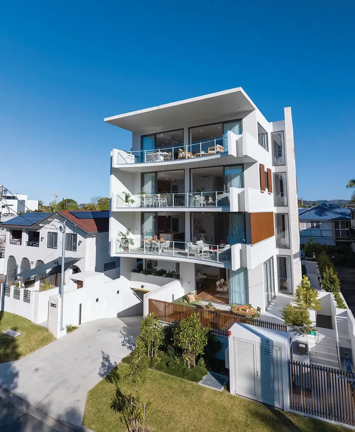 Modern white multi-story building with balconies, facing a sunny blue sky. Driveway leads to entrance