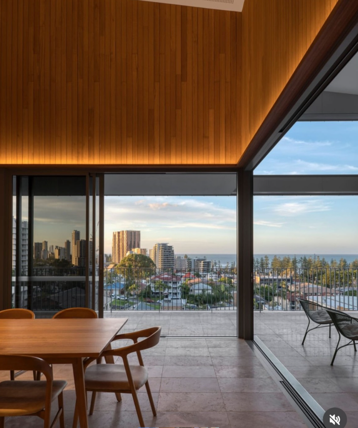 Dining room with wood paneling, open to a balcony with city and ocean views. Table set with chairs.