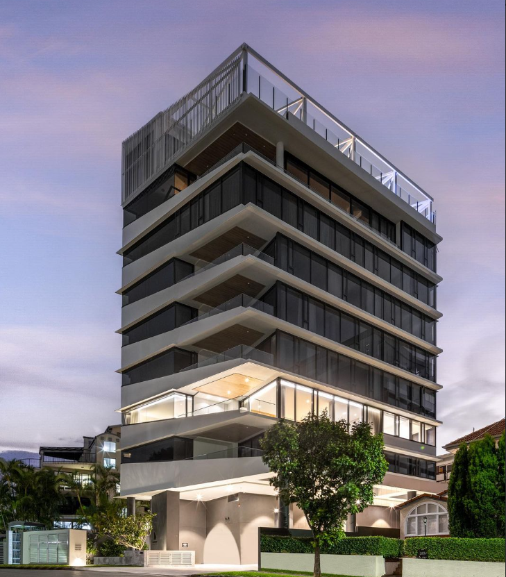 Modern multi-story building with glass windows, white balconies, and a rooftop terrace, set against a dusk sky.