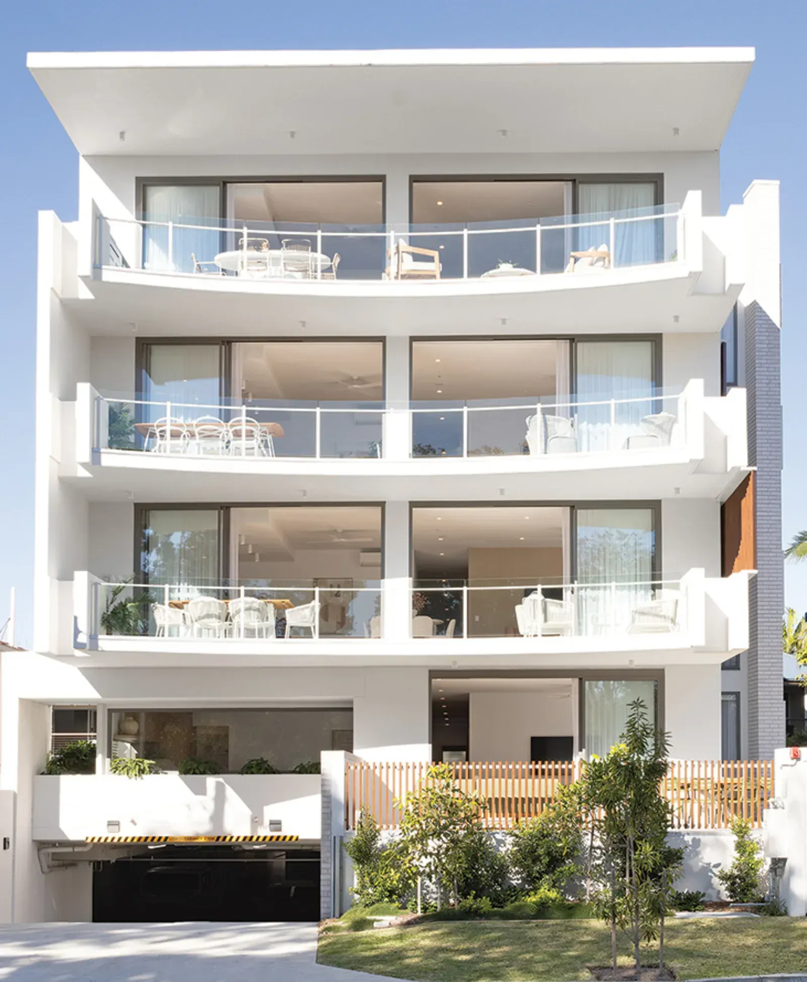 Modern white apartment building with glass balconies, overlooking a lawn and driveway. Black garage door on the ground level.