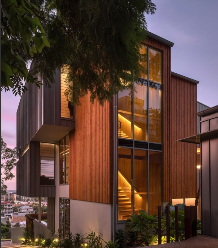 Modern multi-story home with wood and glass exterior, lit stairs visible through large windows, against a dusk sky.