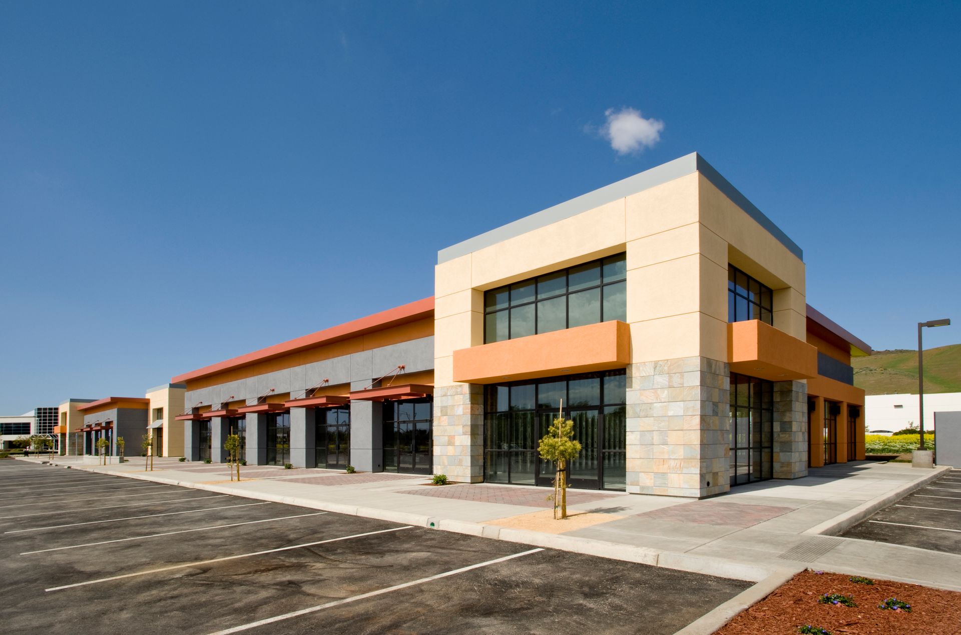 Modern commercial building with orange and tan accents, large windows, and empty parking lot on a sunny day.