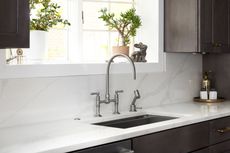 A brushed nickel kitchen faucet with a side sprayer sits on a white countertop against a white marble backsplash.
