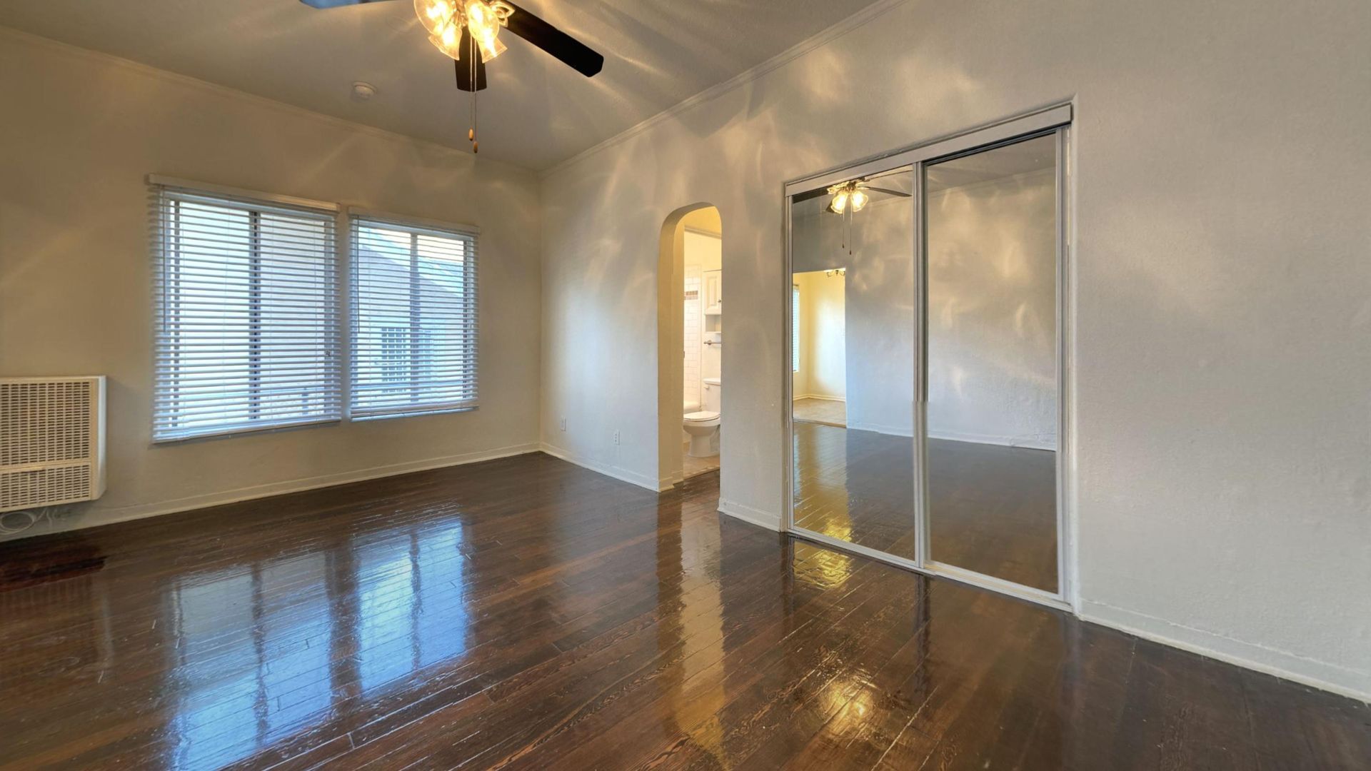 Empty bedroom with dark wood floor, mirrored closet doors, and a window.