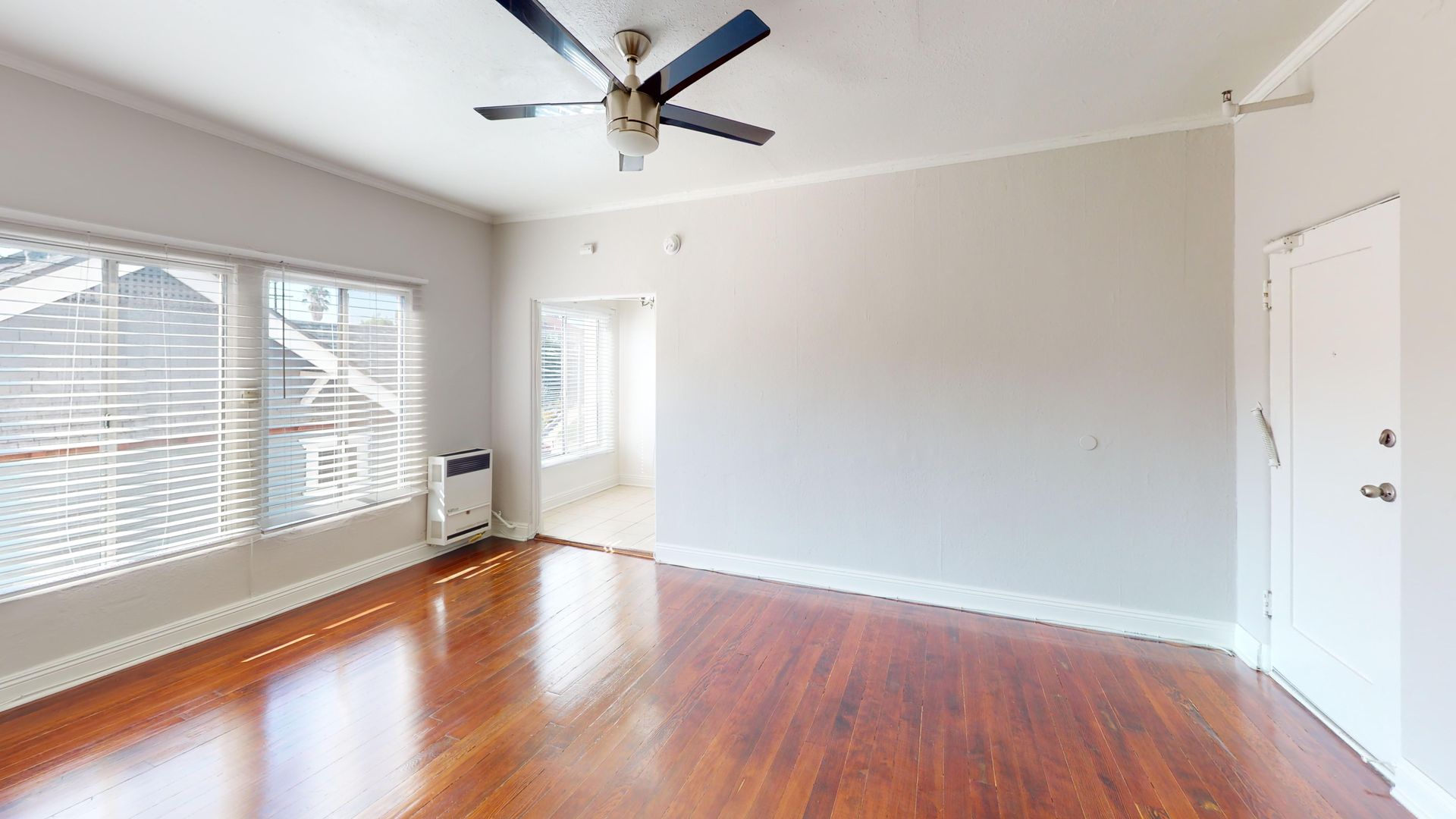 Empty living room with wood floors, large windows, ceiling fan, and white walls.