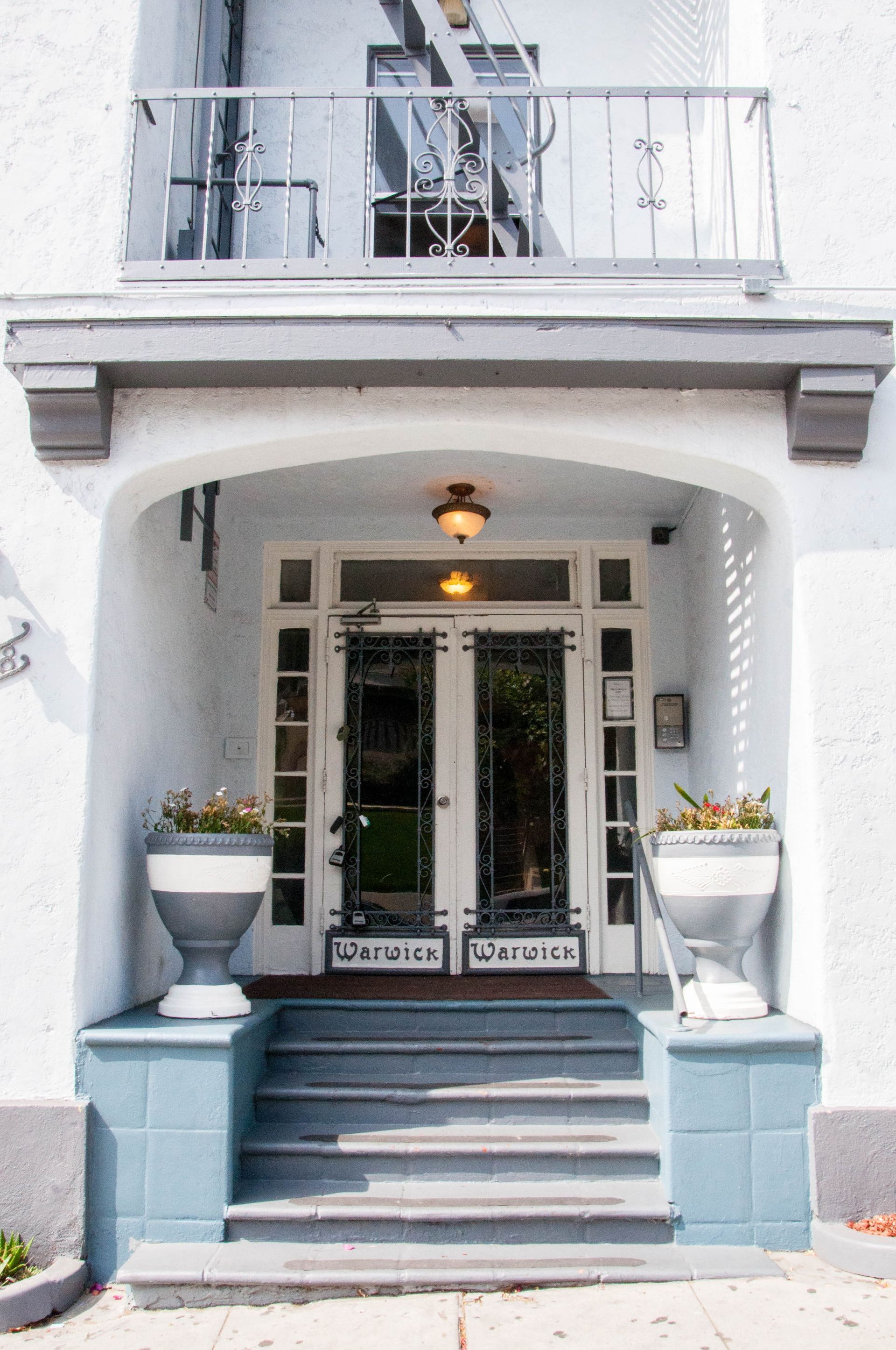 White building entrance with arched doorway, steps, and balcony above. Blue accents, two decorative planters.