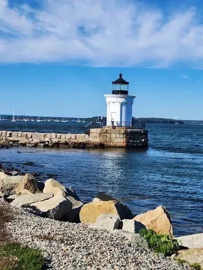 Chimney sweep Portland Maine - A white lighthouse with a black cap stands on a stone pier with sailboats in the distance.