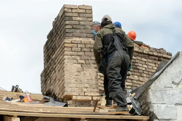 Chimney Sweep Portland Maine - Two workers in safety gear inspect a brick chimney on a roof, surrounded by roofing materials under a cloudy sky.