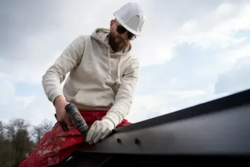 Chimney sweep portland maine - A construction worker uses a power drill to secure metal roofing materials, dressed in a gray sweatshirt and red pants under a cloudy sky.