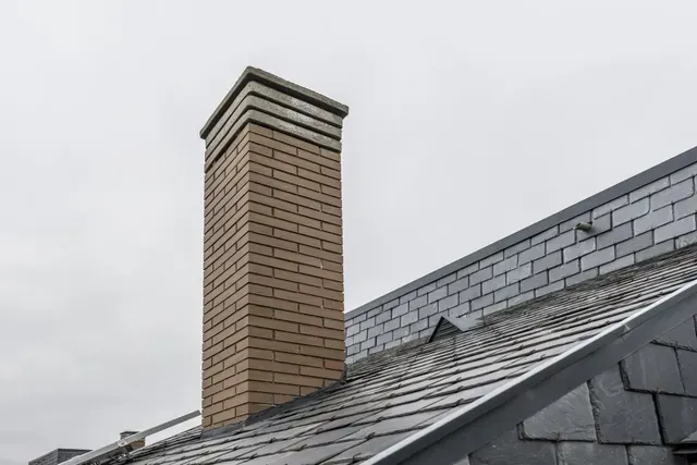 Chimney Sweep Portland Maine - Close-up of a brick chimney atop a slate roof, set against a cloudy sky, showcasing the chimney's rectangular design.