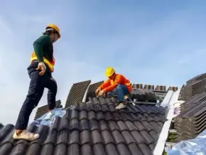 Chimney sweep portland maine - Two roofers wearing hard hats install dark gray tiles on a sloped roof under a clear blue sky.