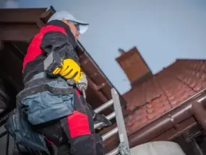 Chimney sweep portland maine - Roofer in yellow gloves and tool belt using a drill while on a ladder next to a brown gutter and red-tiled roof.
