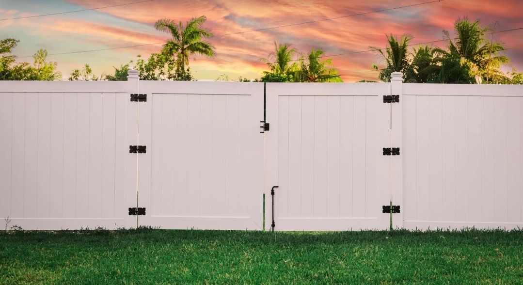 White vinyl fence with closed gate, green grass, and colorful sunset in the background.