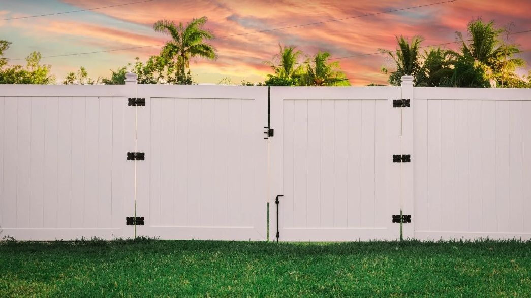 White vinyl fence with closed double gate against a sunset sky and palm trees. Green grass in the foreground.