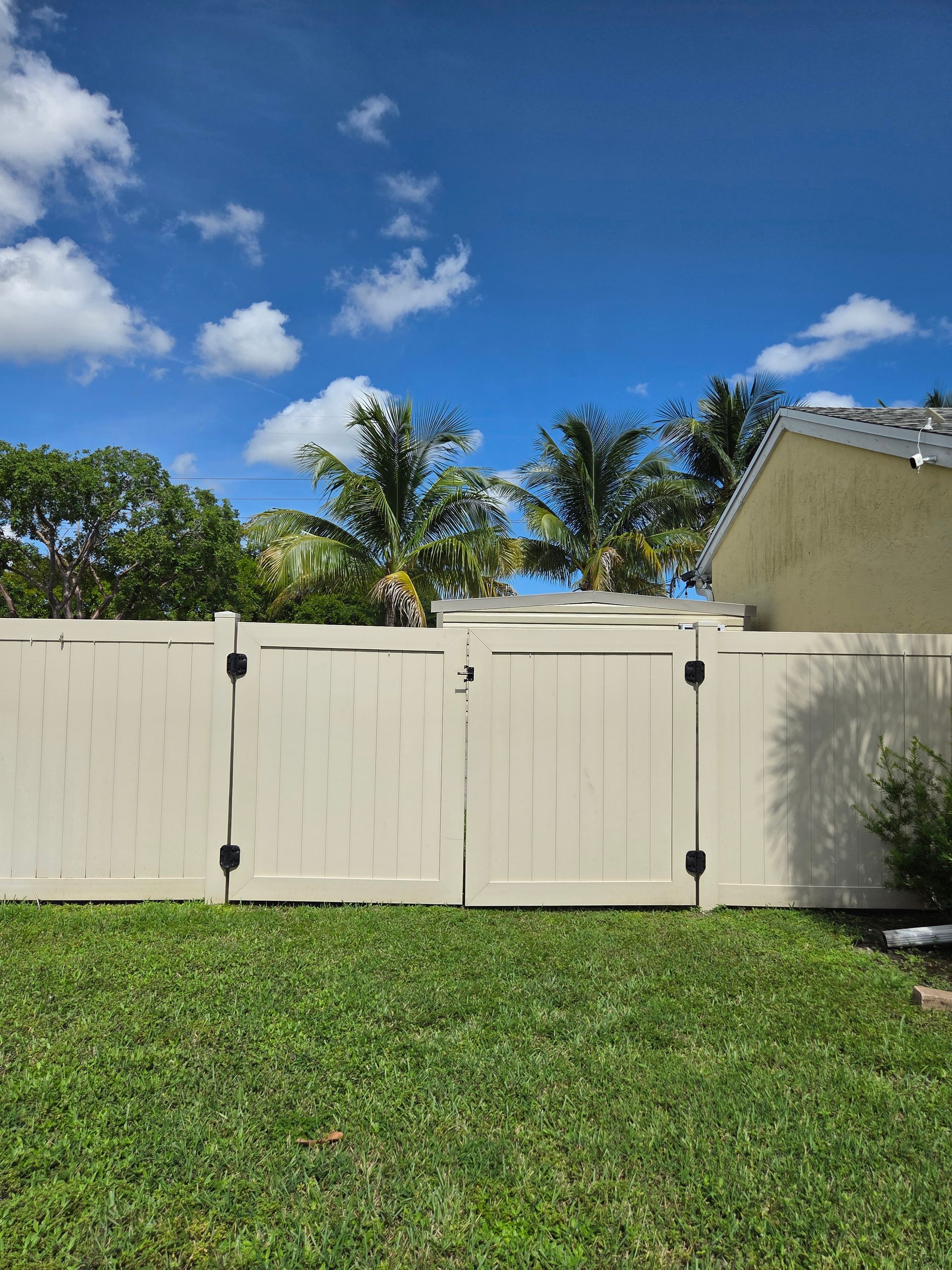 Beige fence with gate in front of green grass, palm trees, and a light blue sky with clouds.