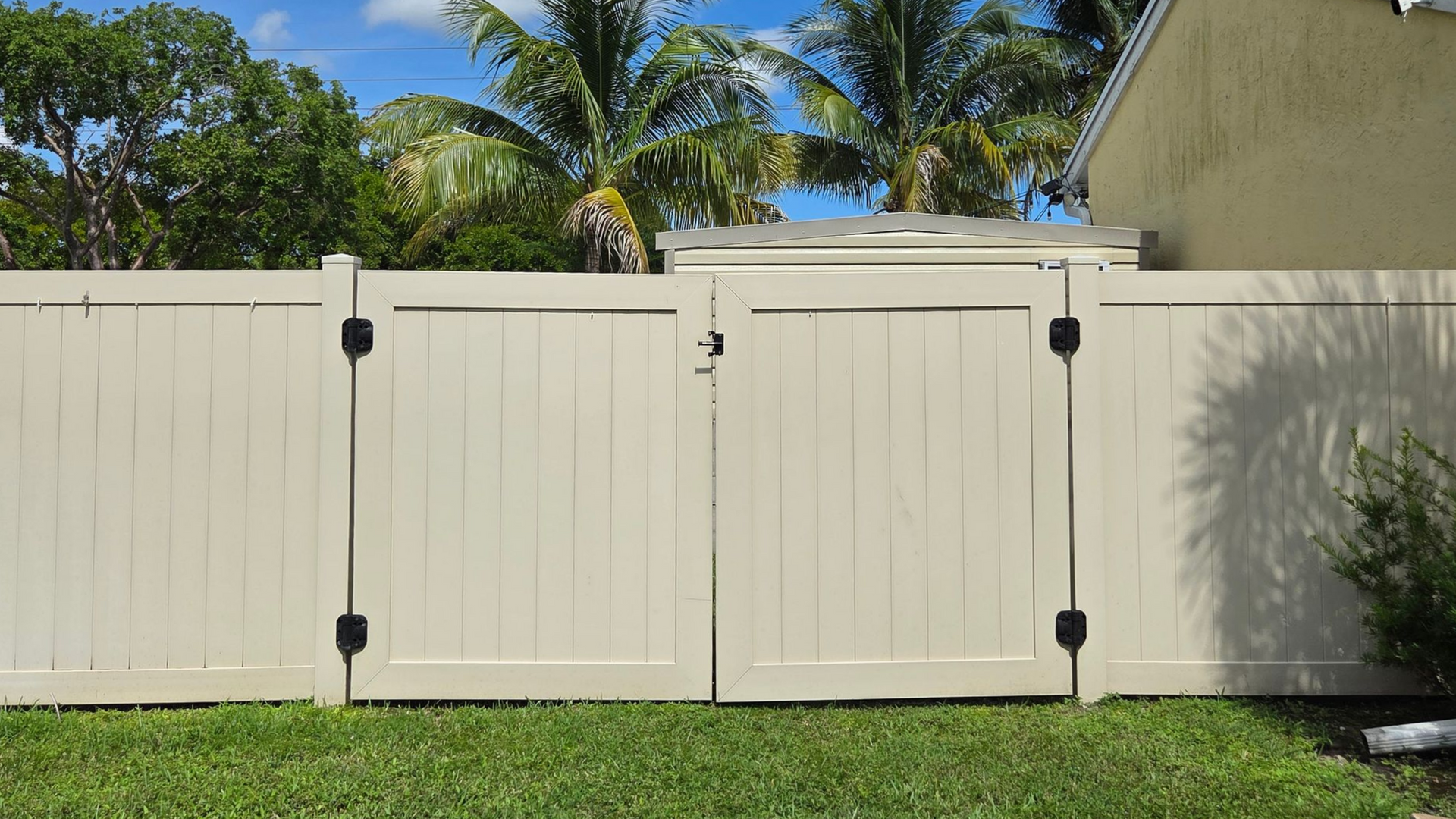 Tan vinyl fence with matching double gate. Black hinges and latch. Green grass and palm trees in background.