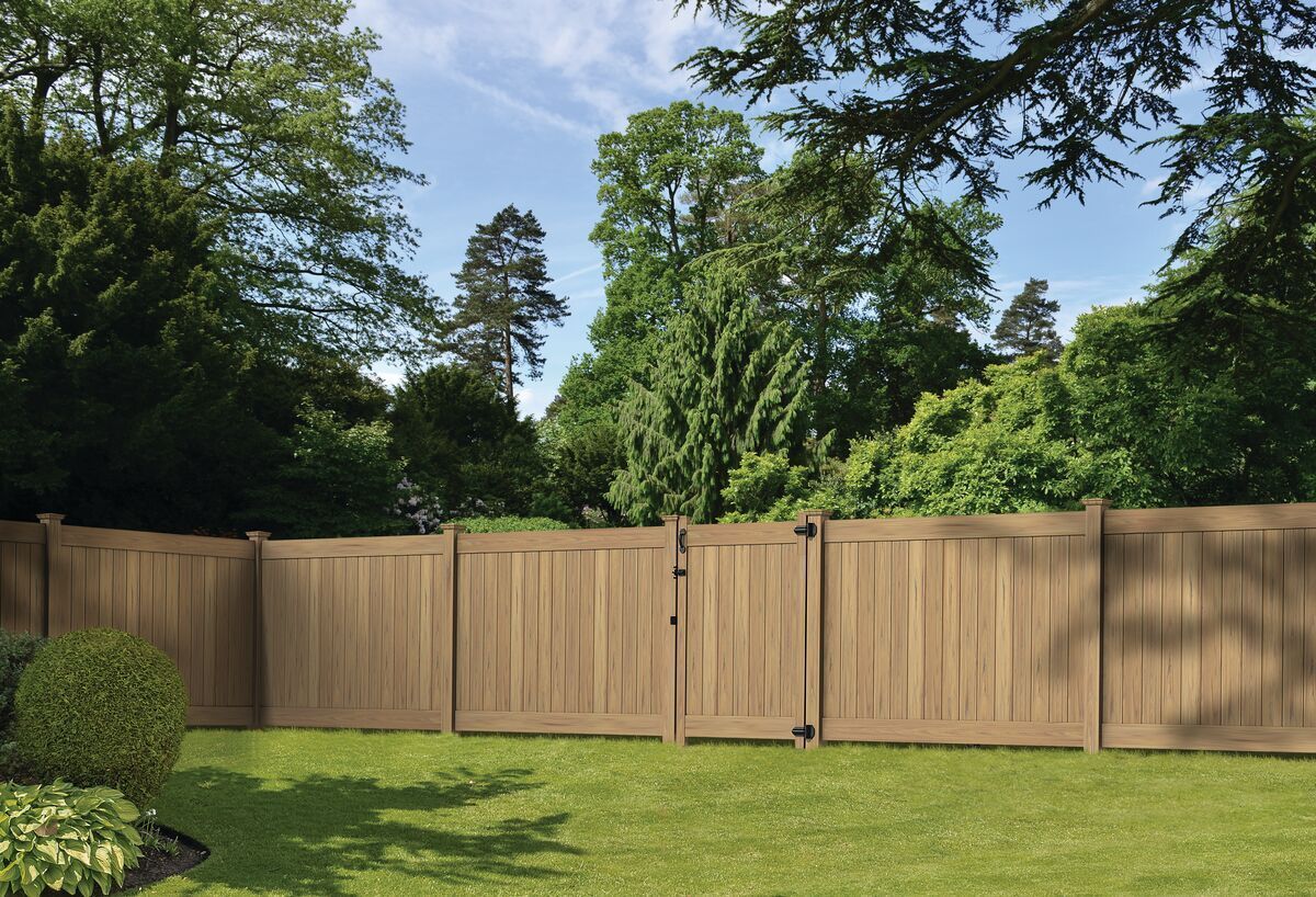Classic wood grain cypress vinyl privacy fence. A wooden fence surrounds a lush green yard with trees in the background.