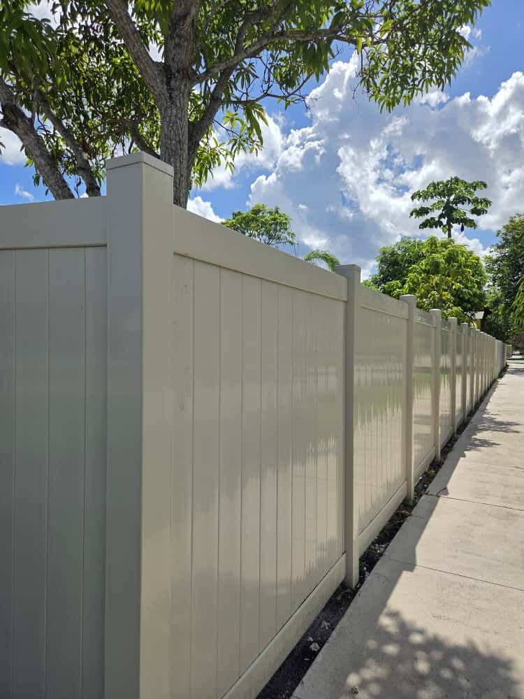 Cream-colored vinyl fence along a sidewalk, under a partly cloudy sky with trees in the background.