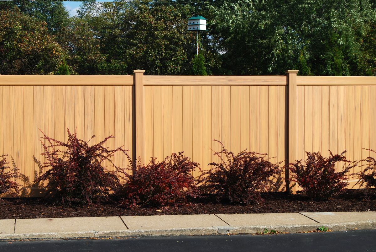 Classic wood grain cypress vinyl privacy fence. A wooden fence with bushes in front of it