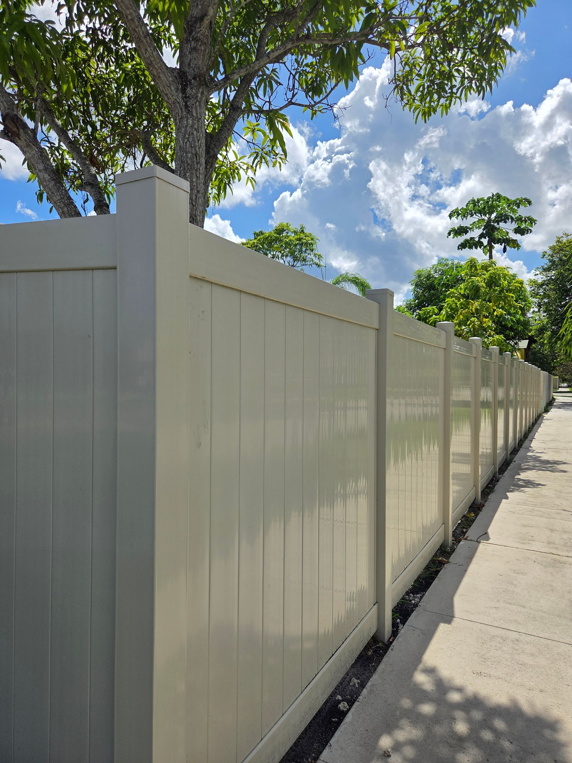 Tan vinyl fence along a sidewalk with blue sky and trees in the background.