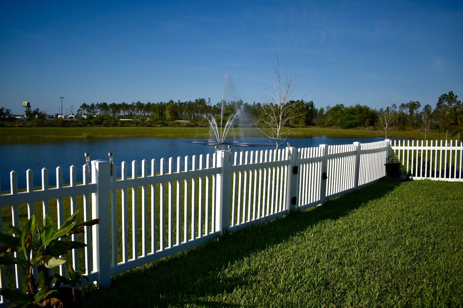 A white picket fence surrounds a pond with a fountain in the background.