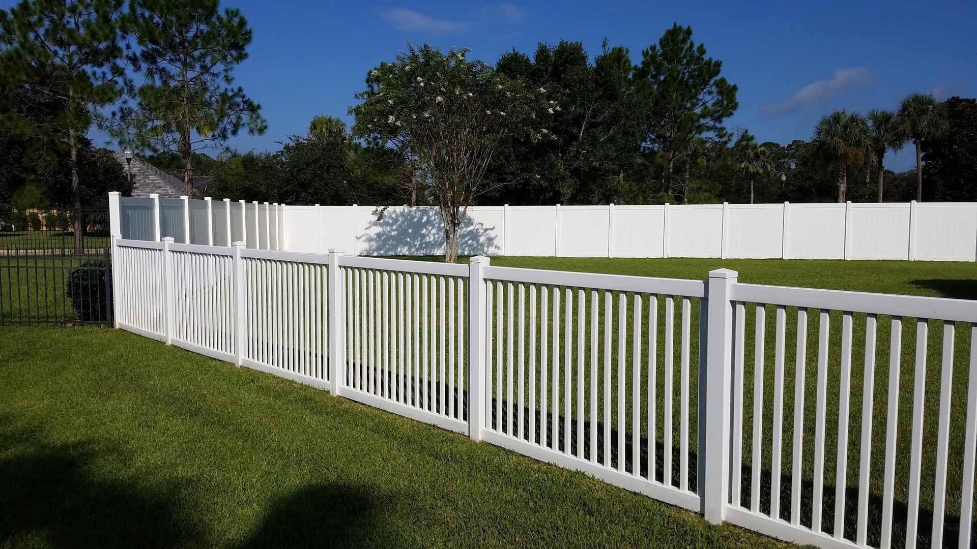 White picket fence in a grassy yard, with a tall white fence in the background against a blue sky.