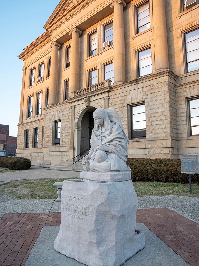 A statue of a woman sitting on a rock in front of a large building.