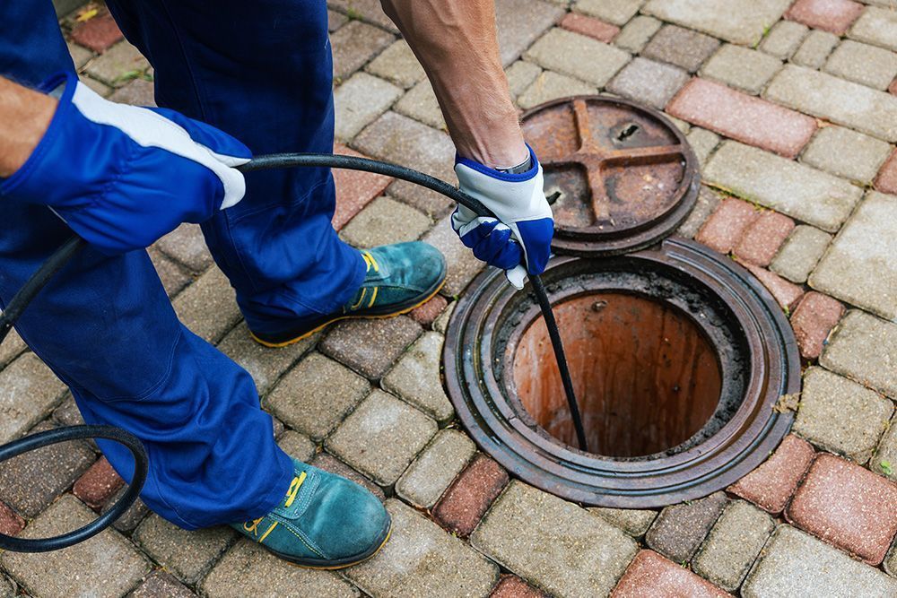 A worker in blue uniform and protective gloves feeds a black cable into an open sewer manhole on a brick-paved surface.