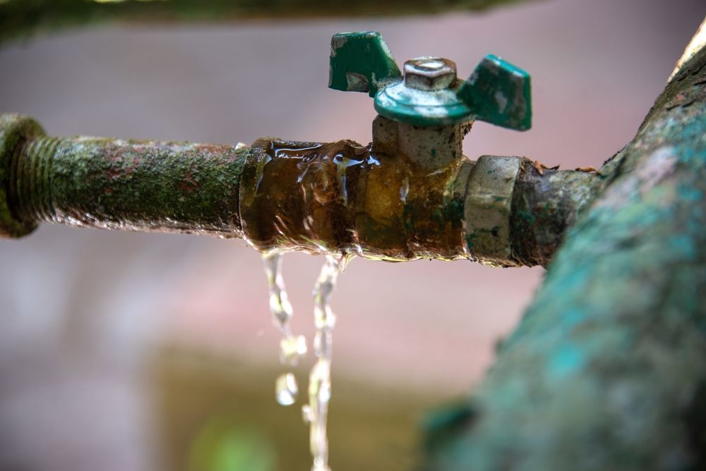 Water leaking from a rusted metal pipe fitting with a green valve handle.