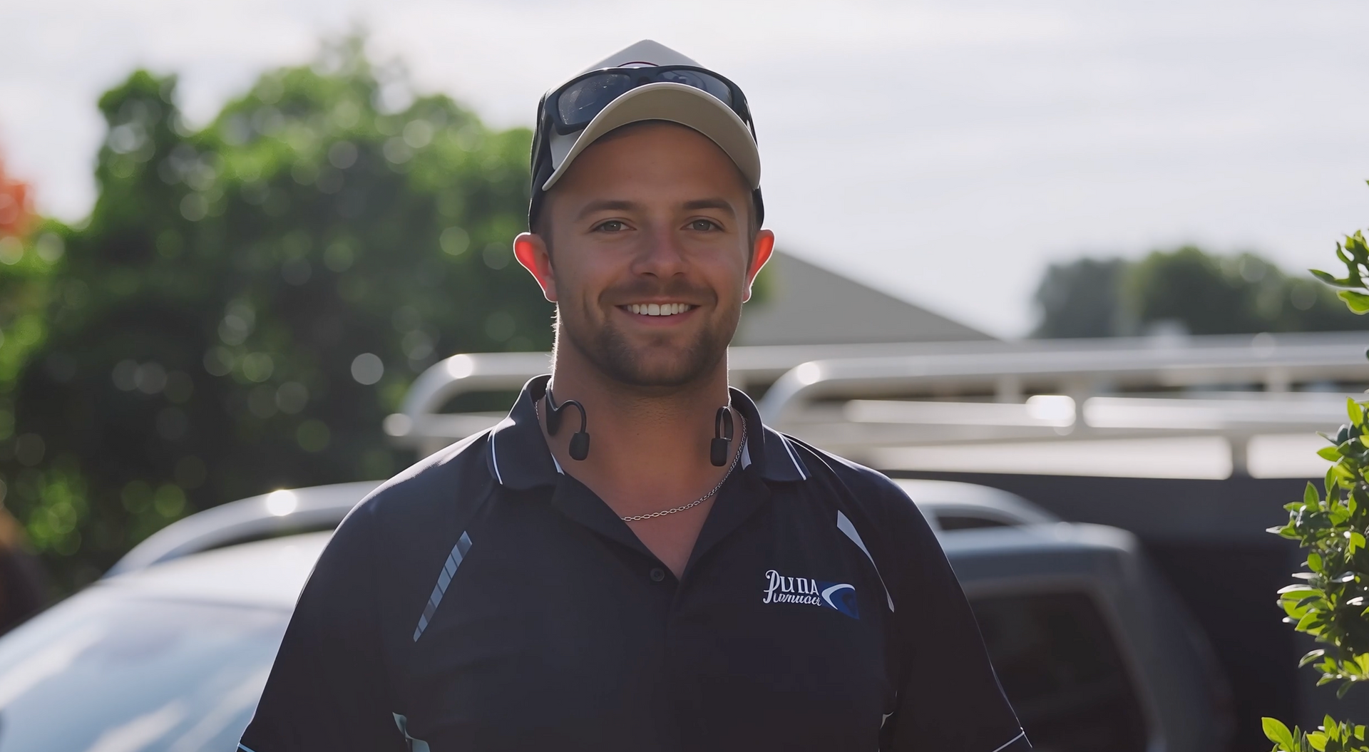 A person wearing a cap, sunglasses, and a dark polo shirt stands outside in front of a vehicle.