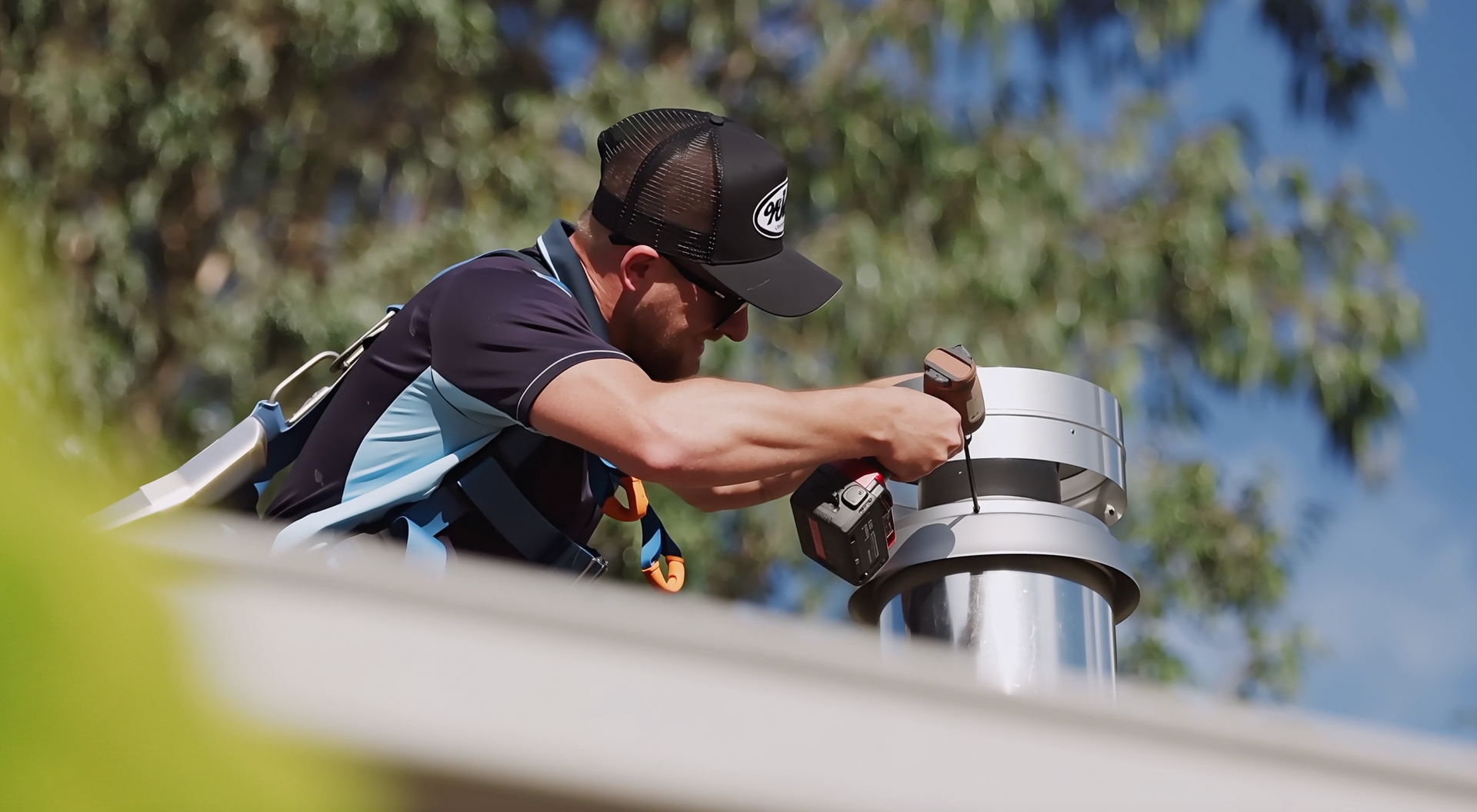 A person wearing a safety harness and cap uses a power drill to install a metal vent pipe on a sloped roof.