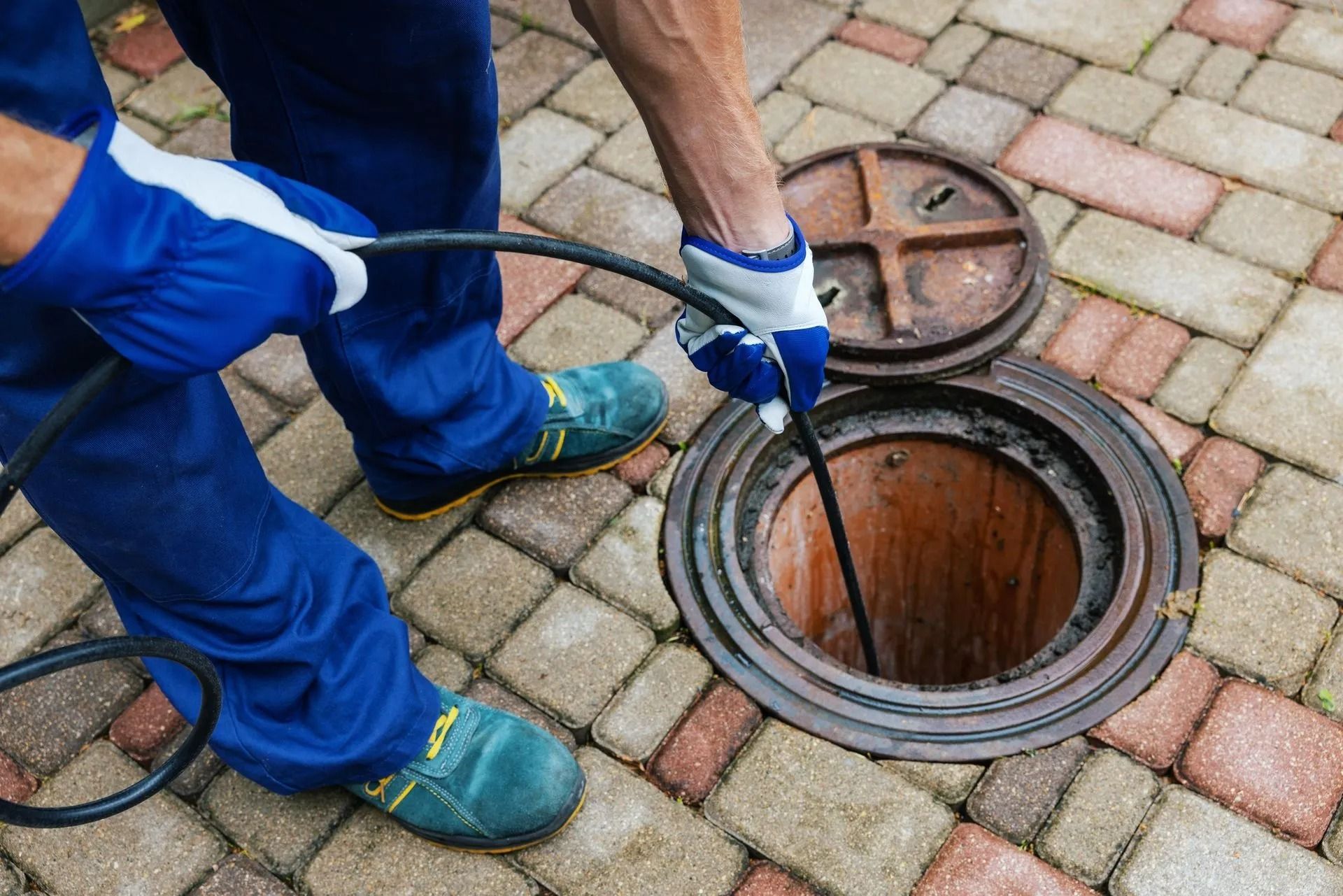 A worker in blue uniform and gloves uses a long cable to inspect or clear an open manhole on a paved surface.
